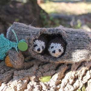 May include: A handmade knitted woodland scene featuring two small hedgehogs nestled inside a gray log. The log is adorned with a green leaf and a small acorn. The scene is set on a textured, brown surface.