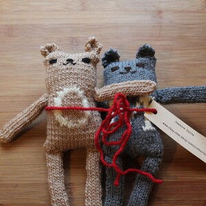 May include: Two knitted bear dolls in tan and gray, with white accents and red yarn details. The dolls are handmade and have a tag attached. The bears are hugging each other, creating a heartwarming scene.