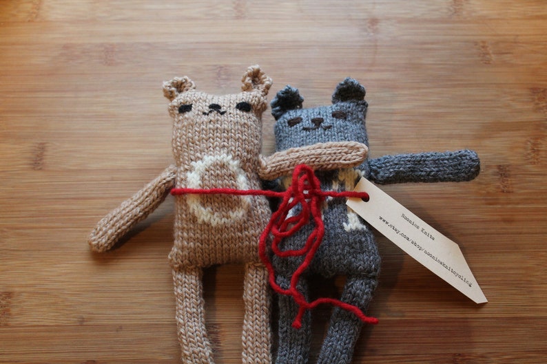 May include: Two knitted bear dolls in tan and gray, with white accents and red yarn details. The dolls are handmade and have a tag attached. The bears are hugging each other, creating a heartwarming scene.