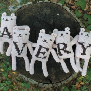 May include: Five handmade, off-white knitted bear dolls spelling out the word "VERY" in brown letters. The bears have small ears, arms, and legs. They are arranged on a weathered tree stump, with green foliage in the background.