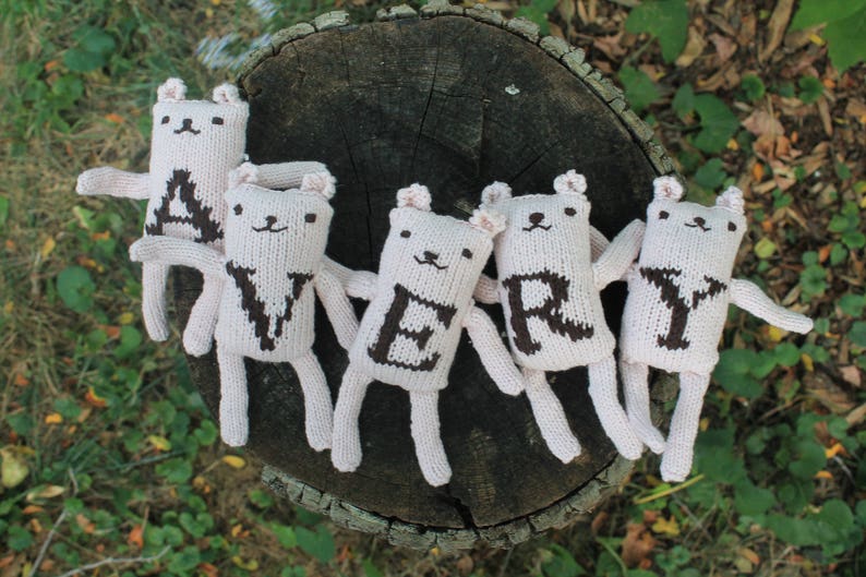 May include: Five handmade, off-white knitted bear dolls spelling out the word "VERY" in brown letters. The bears have small ears, arms, and legs. They are arranged on a weathered tree stump, with green foliage in the background.