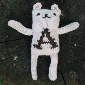 May include: A handmade, light pink knitted bear doll with a dark brown geometric design on its chest. The bear has small ears, a simple face, and outstretched arms and legs. The doll is placed on a dark, textured wooden surface.
