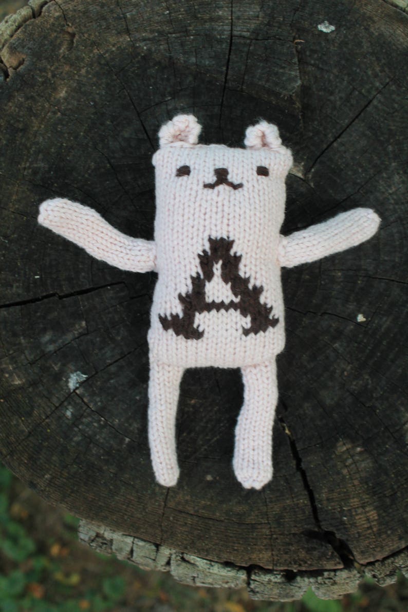 May include: A handmade, light pink knitted bear doll with a dark brown geometric design on its chest. The bear has small ears, a simple face, and outstretched arms and legs. The doll is placed on a dark, textured wooden surface.