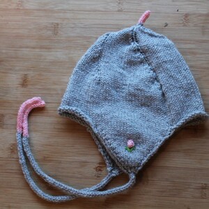May include: A gray knit baby hat with ear flaps and pink accents. The hat features a small pink flower and a pink tip on top. The ties are gray with pink tips. The hat is made of a soft, knitted material.