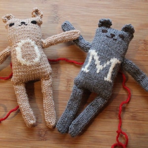 May include: Two handmade knitted bear dolls. One is tan with a white "O" on its chest, the other is gray with a white "M". Both have small black eyes and are connected by a red string.