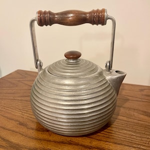 May include: A vintage silver-toned teapot with a wooden handle and lid knob. The teapot has a round body with horizontal ridged detailing. The handle and lid knob are made of dark brown wood. The spout is on the right side of the teapot.