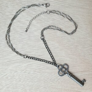 May include: A silver key necklace with a decorative key pendant. The necklace has two chains, one silver and one black.