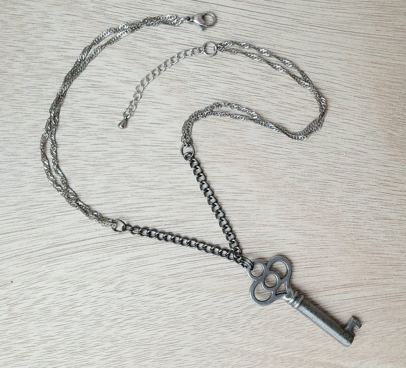 May include: A silver key necklace with a decorative key pendant. The necklace has two chains, one silver and one black.