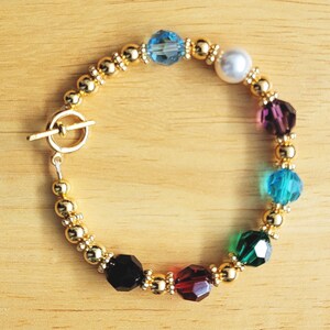 May include: A gold bracelet with a toggle clasp and colorful crystal beads. The beads are blue, white, purple, teal, green, black, and red.