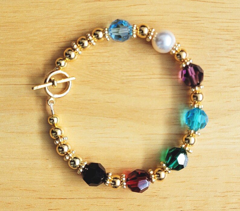 May include: A gold bracelet with a toggle clasp and colorful crystal beads. The beads are blue, white, purple, teal, green, black, and red.