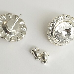 May include: A pair of silver earrings with clear crystal stones. The earrings have a round shape and are surrounded by a row of smaller crystals. The earrings are shown with their backs, which are also silver.