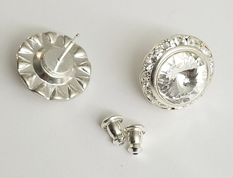 May include: A pair of silver earrings with clear crystal stones. The earrings have a round shape and are surrounded by a row of smaller crystals. The earrings are shown with their backs, which are also silver.