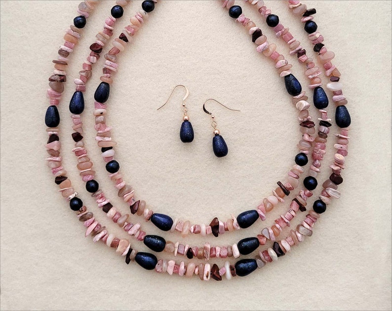 May include: A three-strand necklace made of pink and brown stones with dark blue accents. Matching earrings with dark blue stones hang from gold hooks.