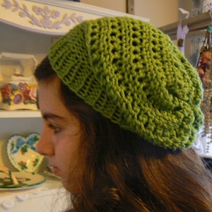 May include: A woman wearing a green crocheted beanie hat. The hat has a lacy design and is pulled down over her forehead.