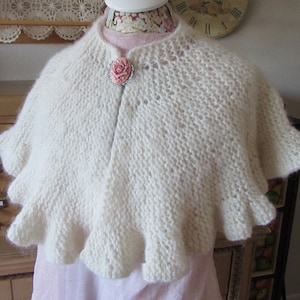 May include: A white knitted shawl with a ruffled edge. The shawl is decorated with a pink flower brooch.