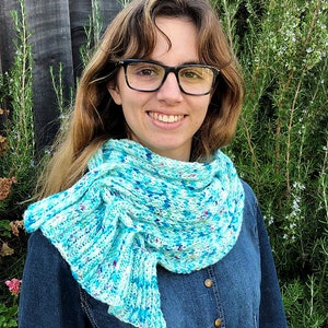 May include: A woman wearing a blue and white knit scarf with a mermaid tail design. The scarf is made with a speckled yarn and has a light blue color with white and pink flecks.