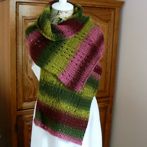 May include: A green, pink, and brown striped knitted scarf with a crocheted design. The scarf is draped over a white dress form.