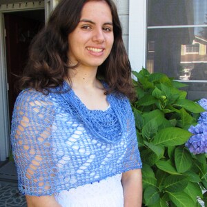 May include: A woman wearing a light blue crocheted shawl with a beaded trim. The shawl has a cowl neck and is made with a delicate openwork stitch. The woman is standing in front of a window with green plants.