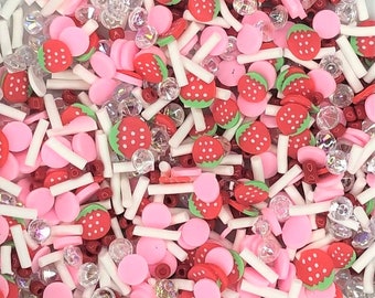 Pode incluir: Uma mistura de confeitos rosa, branco e vermelho, incluindo formas de morango, pequenas contas redondas e palitos brancos longos.