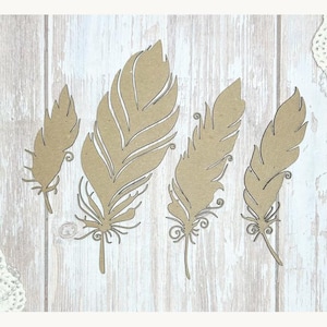 May include: Four decorative feather cutouts in varying sizes and shapes. The feathers are a light brown color and have intricate details, including delicate lines and swirls. The feathers are arranged on a light wood-grain background.