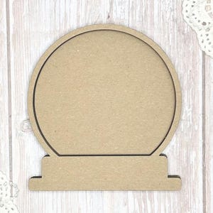 May include: A blank, unfinished wooden snow globe shape. The snow globe has a circular top and a rectangular base. The wood is a light brown color, and the edges are cleanly cut. Perfect for DIY crafts and personalized decorations.