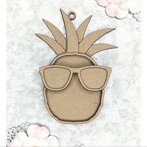 Puede incluir: Adorno de madera con forma de piña con gafas de sol. El adorno es de color marrón claro con una superficie texturizada y un pequeño agujero en la parte superior para colgarlo. Las gafas de sol son rectangulares. Una divertida decoración veraniega.