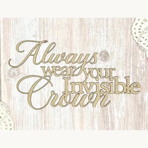 May include: A light brown wooden sign featuring the quote "Always wear your Invisible Crown" in a mix of cursive and block lettering. The sign is set against a light wood-grain background.