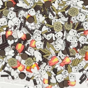 May include: A mix of polymer clay sprinkles in various shapes and colors. The sprinkles include white marshmallows with smiling faces, brown sticks, cookies, hearts, and flames. Small, clear, shiny embellishments are also scattered throughout.