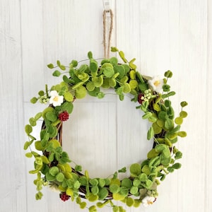 May include: A green felt wreath with white flowers and red berries. The wreath is hanging from a white wall.