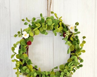 Pode incluir: Uma coroa de feltro verde com flores brancas e bagas vermelhas. A coroa está pendurada em uma parede branca.