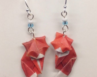 Può includere: Un paio di orecchini a forma di volpe origami in carta rossa e bianca, appesi a ganci d'argento con perline blu e bianche.