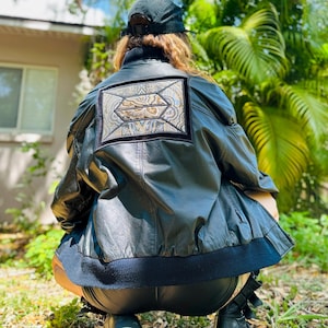 May include: Black leather jacket with a decorative patch on the back. The patch features an ornate design with gold and brown accents. The jacket has a ribbed hem and cuffs. The person is wearing a black cap and black boots.