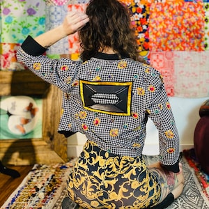 May include: A black and white houndstooth jacket with jewel embellishments and a black leather patch. The jacket is paired with black and gold patterned shorts. The outfit is worn on a patterned rug.