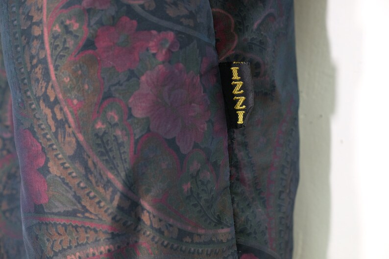 May include: A black fabric with a paisley pattern in shades of pink, green, and brown. The fabric has a black label with the word "IZZI" in gold lettering.
