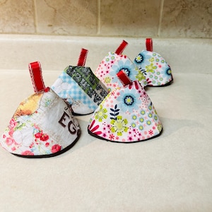 May include: Four cone-shaped fabric tea cozies featuring various patterns including floral prints in pink, blue, and green, and a text print with red roses. Each cozy has a small red ribbon loop at the top and a dark brown fabric base trim.