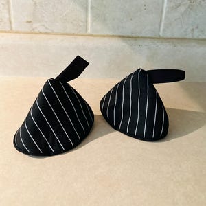 May include: A pair of black oven mitts with a white pinstripe pattern. Each mitt features a conical shape with a rounded base and a black fabric loop attached to the top for hanging. The fabric has a smooth texture and a consistent striped design.