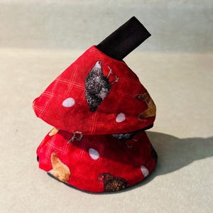 May include: A red fabric pot holder with a plaid pattern featuring printed images of chickens and white eggs. The conical-shaped kitchen accessory includes a black fabric loop at the top for hanging and has a black interior lining visible at the base.