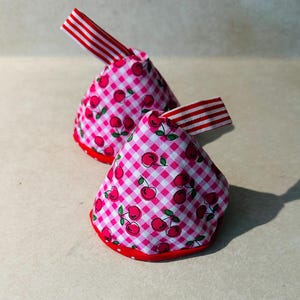 May include: Two cone-shaped fabric tea cozies featuring a pink and white gingham pattern with a red cherry print. Each cozy has a red fabric base and a red and white striped fabric loop attached to the top for hanging or easy handling.