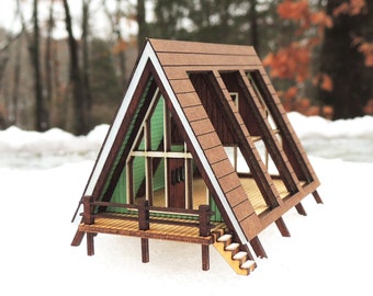 May include: A wooden model of an A-frame cabin with a green and white exterior, a brown wooden deck, and a set of stairs leading up to the front door.