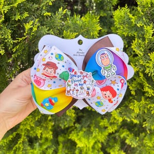 May include: A multi-layered hair bow featuring Toy Story character prints on white, yellow, and rainbow-striped ribbon. The center displays a circular emblem with the text 'You've got a friend in me' and various character illustrations, plus a small blue rocket charm.