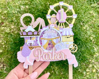 May include: A multi-layered carnival-themed cake topper featuring a Ferris wheel with the number 1, a carousel, a popcorn stand, and a roller coaster silhouette. The design includes a shaker element labelled Parker's Carnival and a pink banner with the name Parker in gold script, all mounted on two wooden sticks.