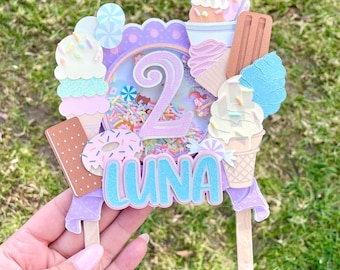 May include: A purple and pink cake topper with a number 2, a donut, ice cream cones, and the name "LUNA" in blue.