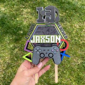 May include: A black and green cake topper with a video game controller, a headset, and the text "Level 13 Reached" and "Jaxson".