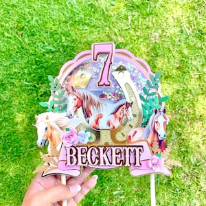 May include: A pink and gold cake topper with a horseshoe, horses, and the number 7. The topper also has the name Beckett on a pink banner.