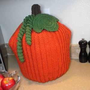 May include: A crocheted pumpkin-shaped cover for an Instant Pot. The cover is orange with a green stem and leaf.