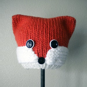 May include: A red and white knitted fox hat with black button eyes and a black button nose. The hat is on a black stand. Woodland Fox Knit Hat.