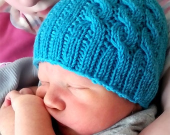 May include: A baby wearing a blue knitted hat with a cable knit pattern. The hat is called "The Baby Asthore Hat".