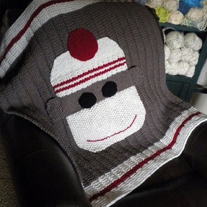 May include: A knitted baby blanket featuring a sock monkey design. The blanket is primarily gray with white, red, and black accents. The sock monkey face is prominently displayed, with a white face, red hat, and black eyes. The text "sock monkey knitted baby blanket" is at the bottom.