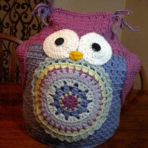 May include: A crocheted owl-shaped instant pot cover in shades of purple, blue, white, and yellow. The owl has large white eyes with black buttons for pupils and a yellow beak.