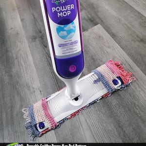 May include: A Swiffer Power Mop with a reusable, crocheted pad in shades of pink, blue, and cream. The mop handle is white and purple, with the Swiffer logo and product information visible. The image also includes the text "Reusable Swiffer Power Mop Pad Pattern".